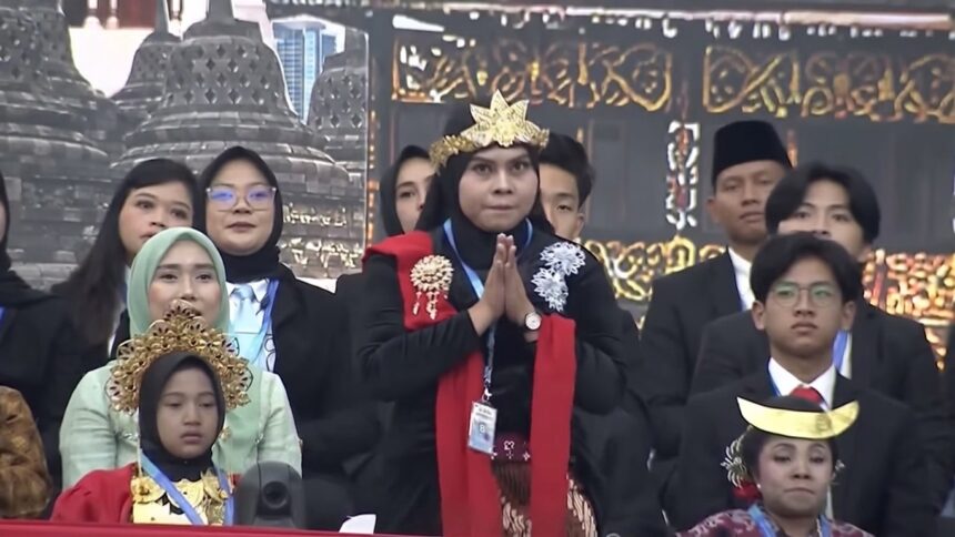 Person in black attire with a golden crown and red scarf stands with hands pressed together in a prayer pose on a stage, surrounded by formally dressed attendees.