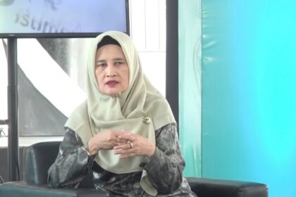 Woman in a beige hijab and patterned gray blouse seated on a dark sofa, hands clasped, speaking at an event.