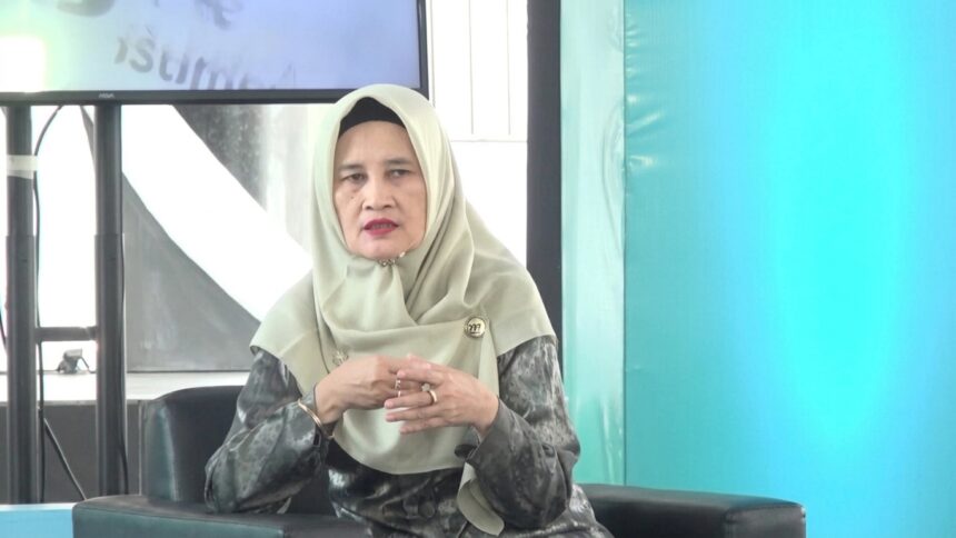 Woman in a beige hijab and patterned gray blouse seated on a dark sofa, hands clasped, speaking at an event.