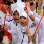 A group of children in white uniforms waving red flags, celebrating together at a crowd event.