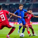 Soccer players in blue and red uniforms contest the ball on a green pitch during a match.