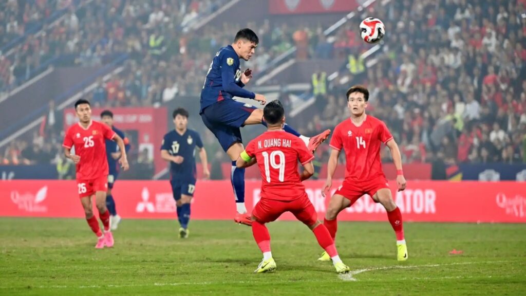 Soccer match scene: a blue-jersey player leaps to kick the ball while red-jersey defenders look on on a crowded stadium pitch.