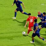 Red team player wearing number 18 dribbles the ball past blue-clad defenders on a bright green pitch.
