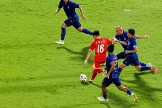 Red team player wearing number 18 dribbles the ball past blue-clad defenders on a bright green pitch.