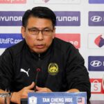 Press conference speaker in a black jacket with crest, seated at a table with a microphone and nameplate reading 'TAN CHENG HOE'.
