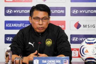Press conference speaker in a black jacket with crest, seated at a table with a microphone and nameplate reading 'TAN CHENG HOE'.