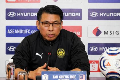 Press conference speaker in a black jacket with crest, seated at a table with a microphone and nameplate reading 'TAN CHENG HOE'.