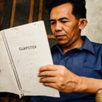 Man in a blue shirt sits reading a worn, white booklet labeled 'GLAPOTEK' held close for focus.