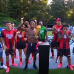 Shirtless young man raises a gold trophy while teammates in red-and-black uniforms celebrate on a soccer field.
