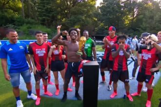 Shirtless young man raises a gold trophy while teammates in red-and-black uniforms celebrate on a soccer field.