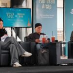 Panel discussion at a book fair: three speakers on gray couches, papers in hand, with drinks on the table and turquoise banners behind reading Jogja Book Fair 2026 and Pameran Arsip.