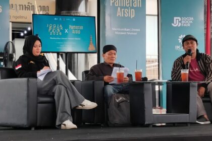 Panel discussion at a book fair: three speakers on gray couches, papers in hand, with drinks on the table and turquoise banners behind reading Jogja Book Fair 2026 and Pameran Arsip.