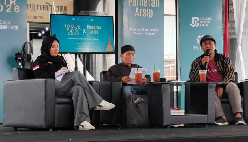 Panel discussion at a book fair: three speakers on gray couches, papers in hand, with drinks on the table and turquoise banners behind reading Jogja Book Fair 2026 and Pameran Arsip.
