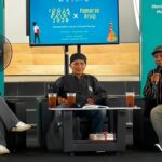 Panel discussion with three speakers seated on stage, each with drinks on a small table, teal backdrop banners behind them.