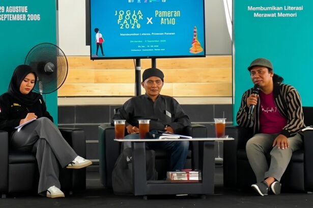 Panel discussion with three speakers seated on stage, each with drinks on a small table, teal backdrop banners behind them.