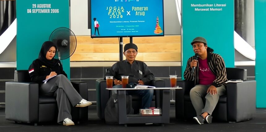 Panel discussion with three speakers seated on stage, each with drinks on a small table, teal backdrop banners behind them.
