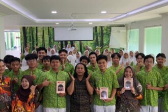 Group of students in matching green shirts posing for a photo in a classroom, two adults in front holding books/destinations, forest mural in the background.