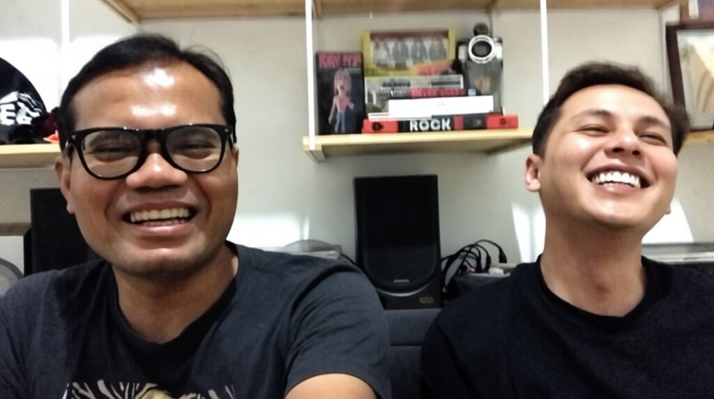 Two men smiling for a selfie in a room with shelves, books, and audio gear (speaker) in the background; one wears glasses on the left.