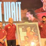 Group of people in maroon polo shirts standing on a stage with a framed poster board featuring signed photos of athletes.