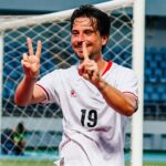 Soccer player in a white jersey with number 19, smiling and making two victory fingers in front of a goal net.