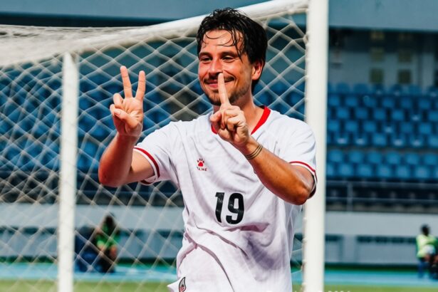 Soccer player in a white jersey with number 19, smiling and making two victory fingers in front of a goal net.