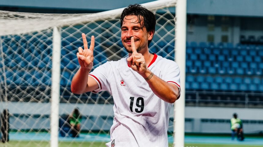 Soccer player in a white jersey with number 19, smiling and making two victory fingers in front of a goal net.