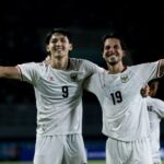 Two soccer players in white jerseys (numbers 9 and 19) celebrate with arms around each other on a night field, smiles on their faces.