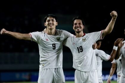 Two soccer players in white jerseys (numbers 9 and 19) celebrate with arms around each other on a night field, smiles on their faces.