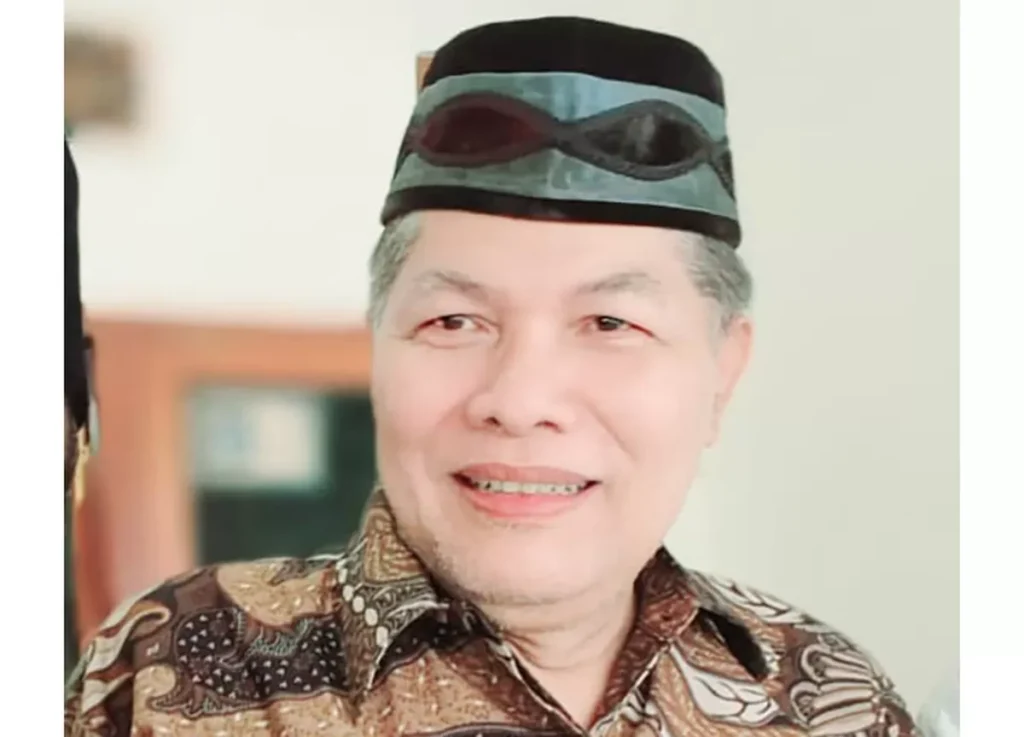Smiling middle-aged man in a batik shirt, wearing a black cap with a blue-green band and sunglasses on top of the cap