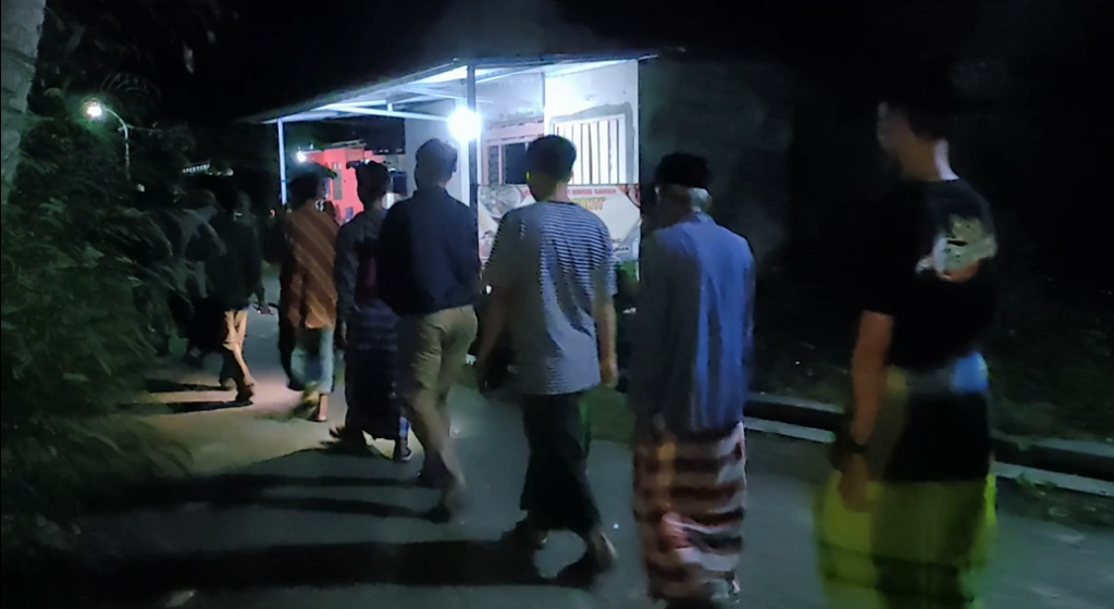 Untitled1 - Mabur.co People walk in a line at night toward a brightly lit food stall or kiosk along a dark path, with foliage to the left and a railing to the right.