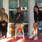 Three young women sit on stools on a red carpet stage; center woman speaks into a microphone while the others listen.