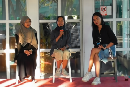 Three young women sit on stools on a red carpet stage; center woman speaks into a microphone while the others listen.