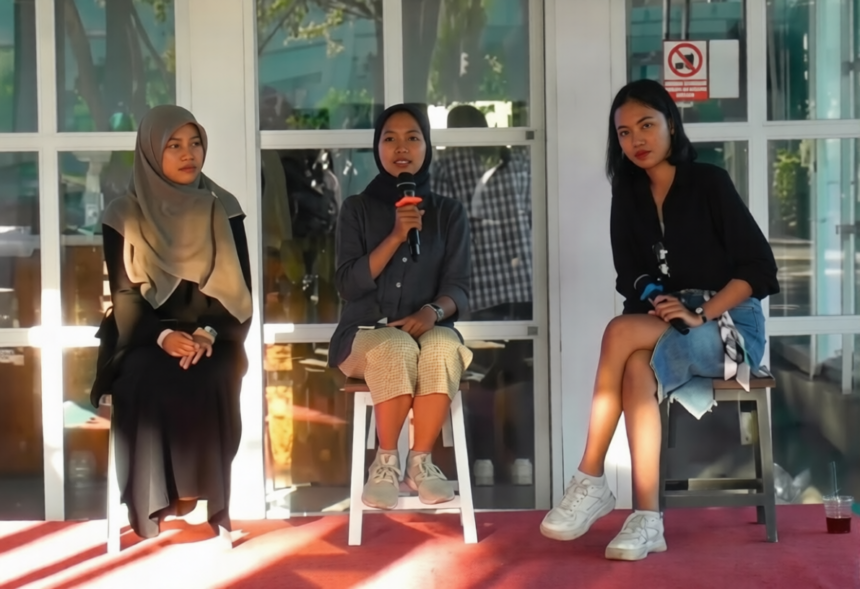 Three young women sit on stools on a red carpet stage; center woman speaks into a microphone while the others listen.
