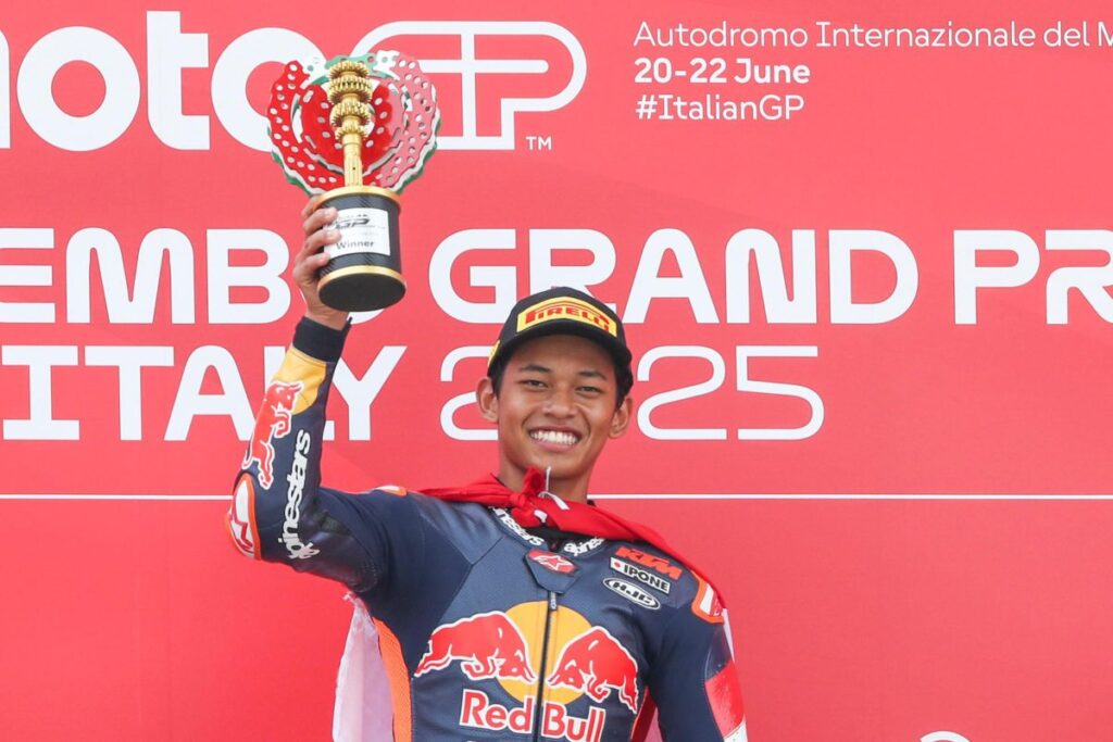VEP9 RBRC ANTARANEWS com - Mabur.co Male MotoGP rider in Red Bull suit on a podium, raising a trophy in front of a red backdrop that reads'Grand Prix Italy 2025'