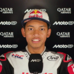 Racer in white racing suit and Red Bull cap being interviewed, with sponsor logos (Qatar Airways, MotoGP) in the background.