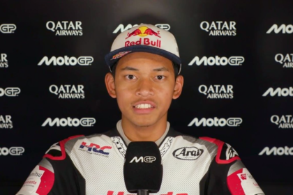 Racer in white racing suit and Red Bull cap being interviewed, with sponsor logos (Qatar Airways, MotoGP) in the background.