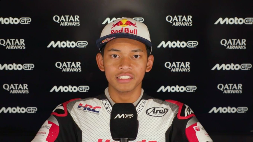 Racer in white racing suit and Red Bull cap being interviewed, with sponsor logos (Qatar Airways, MotoGP) in the background.