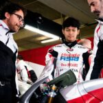 Young rider in a Honda Asia racing suit stands between two team members in a garage, inspecting a red-and-white motorcycle.