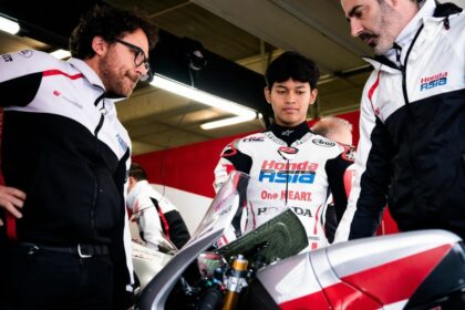Young rider in a Honda Asia racing suit stands between two team members in a garage, inspecting a red-and-white motorcycle.