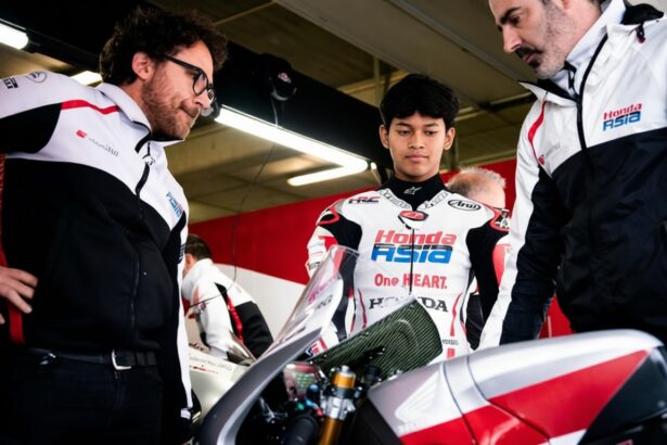 Young rider in a Honda Asia racing suit stands between two team members in a garage, inspecting a red-and-white motorcycle.