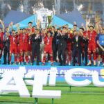 Soccer team in red uniforms celebrates on stage after winning, raising arms with the trophy as fireworks light the stadium.