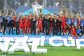 Soccer team in red uniforms celebrates on stage after winning, raising arms with the trophy as fireworks light the stadium.