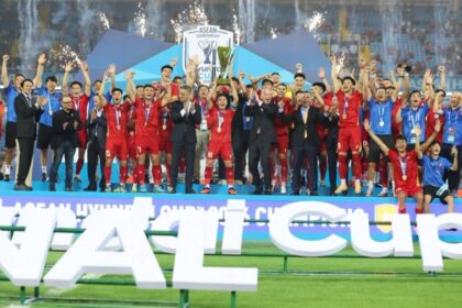 Soccer team in red uniforms celebrates on stage after winning, raising arms with the trophy as fireworks light the stadium.