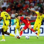 Red-jersey soccer player dribbles the ball between two yellow-jersey defenders on a green pitch with a packed stadium in the background.