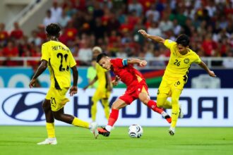 Red-jersey soccer player dribbles the ball between two yellow-jersey defenders on a green pitch with a packed stadium in the background.