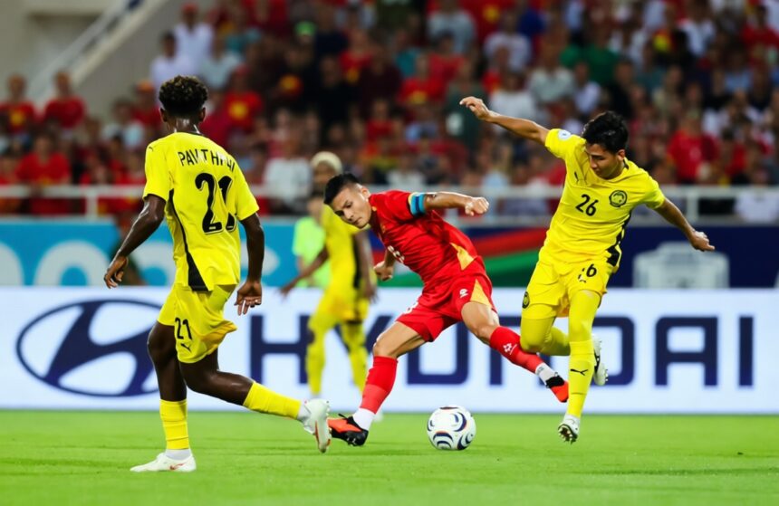 Red-jersey soccer player dribbles the ball between two yellow-jersey defenders on a green pitch with a packed stadium in the background.