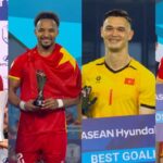 Four male soccer players in red and yellow uniforms pose with trophies on a prize stage against a blue backdrop.