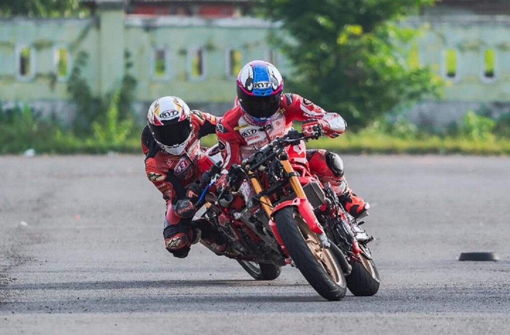 Two motorbike racers in red protective suits lean into a sharp turn on a paved track.