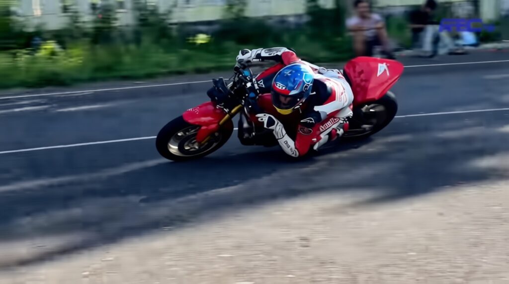 Rider in a red-and-white suit leans into a sharp turn on a red sport motorcycle on a road race course with spectators in the background, blue helmet visible.