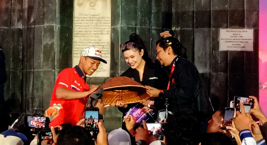Three people on a stage hand a large woven basket to a smiling man in a red shirt as photographers in the crowd capture the moment with their phones.
